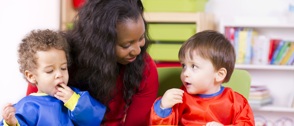 Two toddlers eating fruit sitting at a child's table in a nursery setting. A carer is sitting behind them.