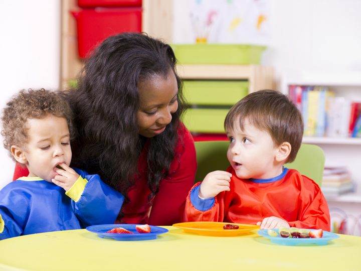 Two toddlers eating fruit sitting at a child's table in a nursery setting. A carer is sitting behind them.