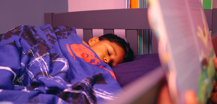 A child sleeping in their bed. Nearby an adult is reading a children's book to them.