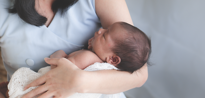 A woman sitting down and smiling whilst looking down at the baby she is holding, which is wrapped in a blanket, in her arms