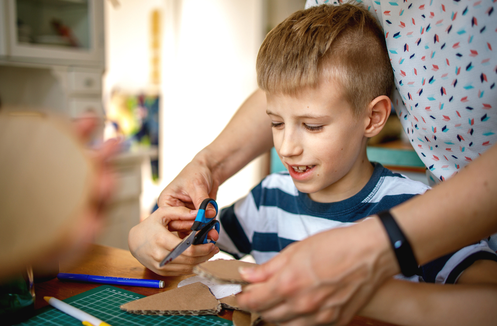 Child sitting at a table with adult supporting their scissor skills.