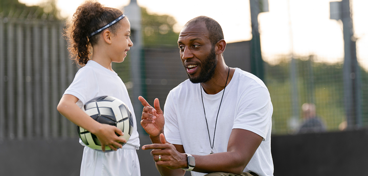 Adult kneeling talking to a child in sportswear holding a ball.