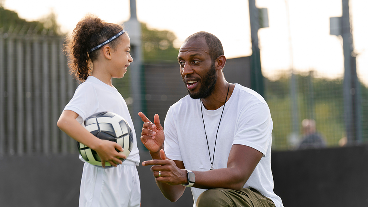 Adult kneeling talking to a child in sportswear holding a ball.