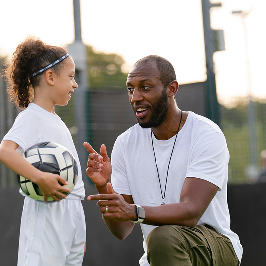 Adult kneeling talking to a child in sportswear holding a ball.