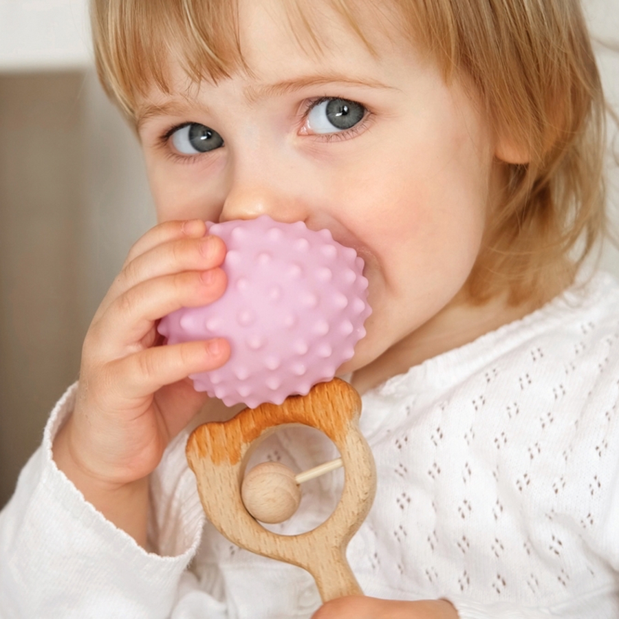 Young girl holding a wooden rattle while chewing on a plastic ball.