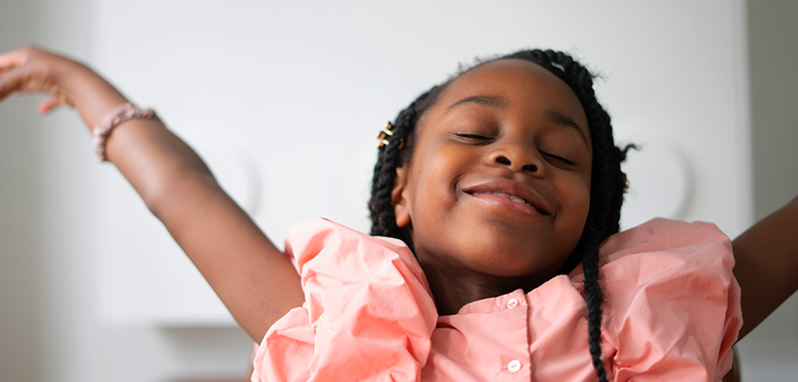 A child smiling with her eyes closed. She's wearing a pink dress with her arms raised above her head gracefully.