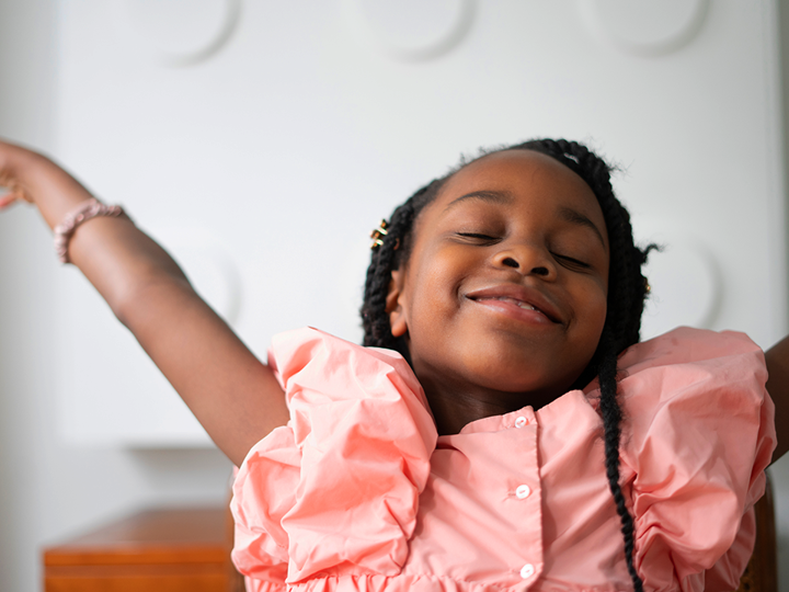 A child smiling with her eyes closed. She's wearing a pink dress with her arms raised above her head gracefully.