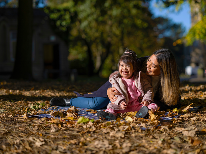 Down's syndrome child and parent laughing and sitting on picnic blanket in autumn with leaves around them.