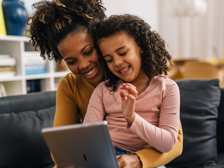 little girl sitting on lap of adult woman on a sofa looking at a handheld tablet device. 