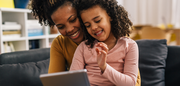 little girl sitting on lap of adult woman on a sofa looking at a handheld tablet device.
