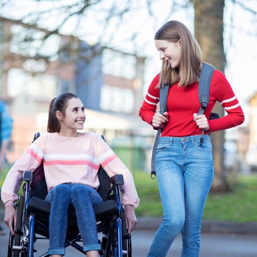 Two young girls talking. One girl is sitting in a wheelchair whilst the other girl is standing next to her with a backpack on.