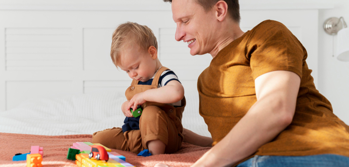 Father and son sitting on the bed. The young boy is playing with coloured blocks and toy cars.