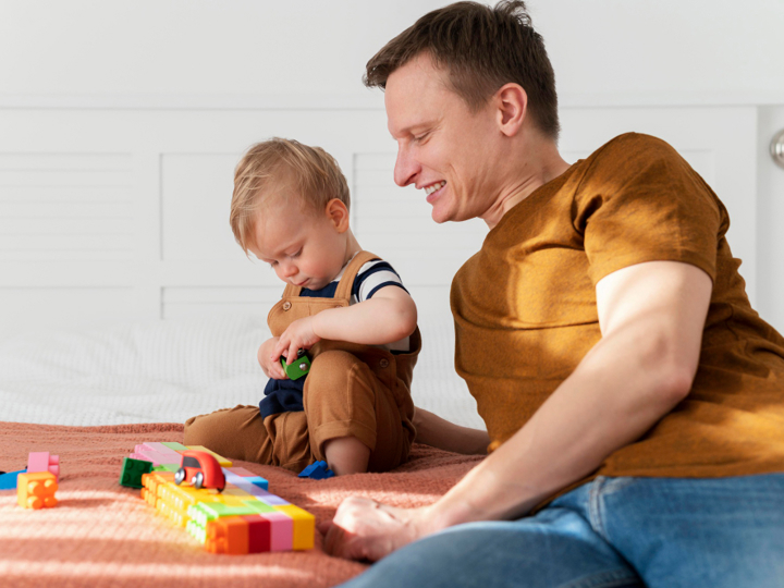Father and son sitting on the bed. The young boy is playing with coloured blocks and toy cars.