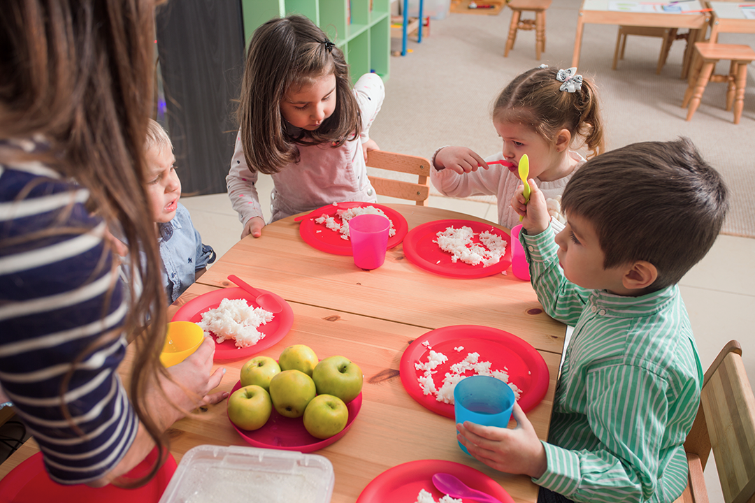 Group of 4 young children sitting round a table eating food from plastic plates.