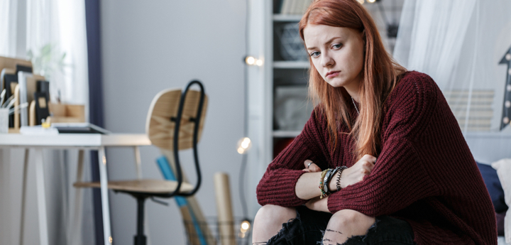 Teenage girl sitting on bed looking upset and concerned with her arms crossed and resting on her knees.