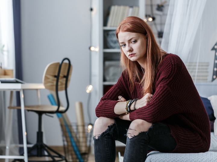 Teenage girl sitting on bed looking upset and concerned with her arms crossed and resting on her knees.