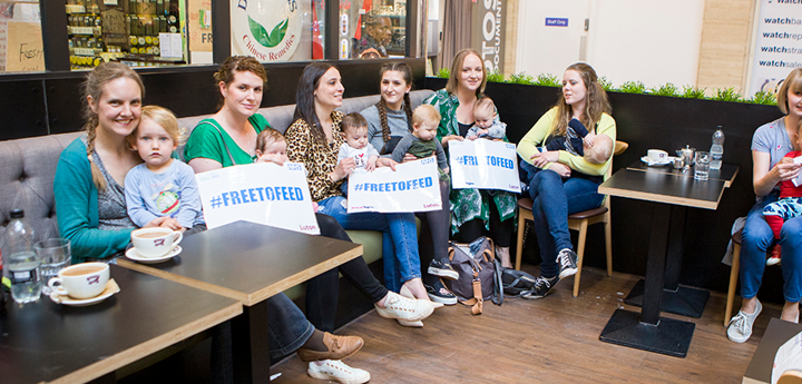 7 mums sitting in a cafe with their babies on their lap. Some of them are holding 'Free to Feed' signs.