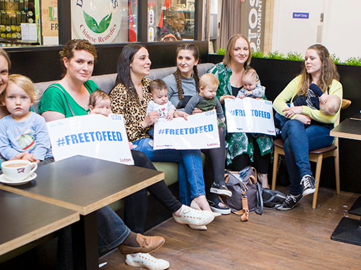 7 mums sitting in a cafe with their babies on their lap. Some of them are holding 'Free to Feed' signs.