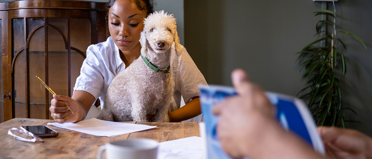 Woman writing at a table with a white dog on her lap. In foreground is a coffee mug and a hand turning a page of a book.