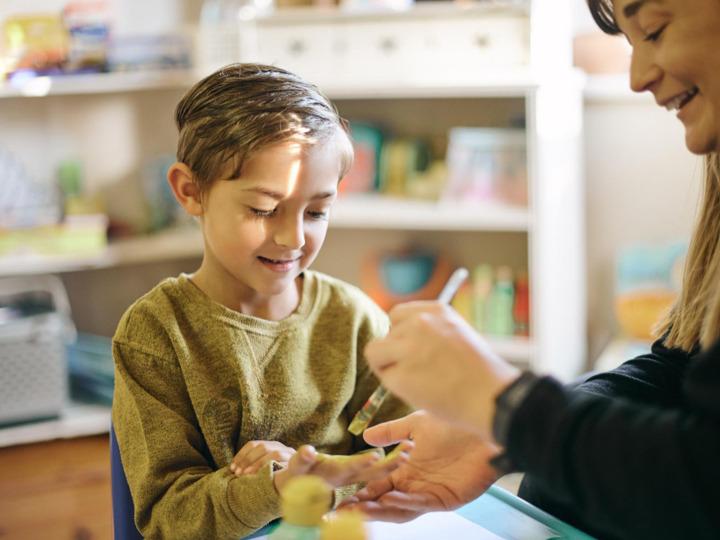 Teacher using a paintbrush to paint a child's hands. The child is smiling and holding out their hand to be painted