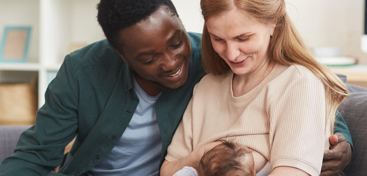 Mum sitting on sofa breastfeeding her baby with the dad sitting next to her with his arm round her. 