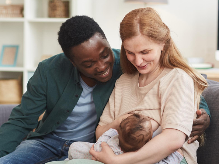 Mum sitting on sofa breastfeeding her baby with the dad sitting next to her with his arm round her. 