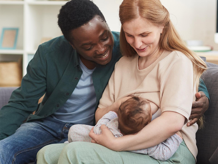 Mum sitting on sofa breastfeeding her baby with the dad sitting next to her with his arm round her. 