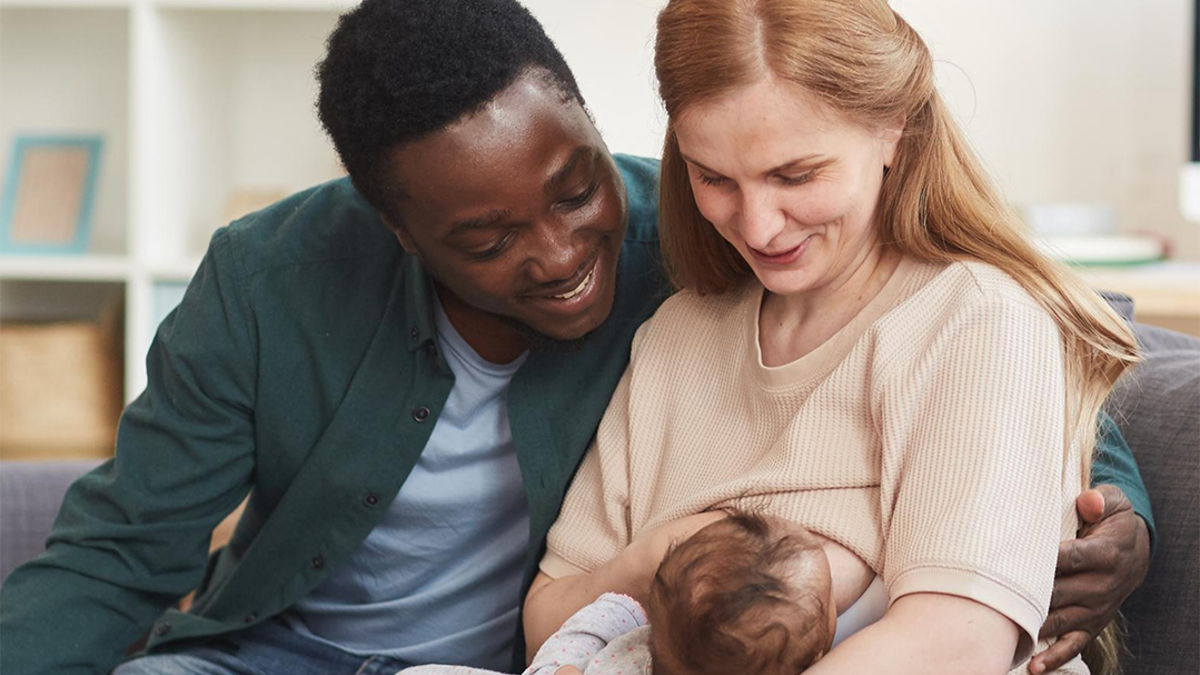 Mum sitting on sofa breastfeeding her baby with the dad sitting next to her with his arm round her. 