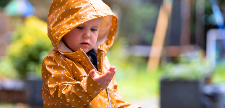 Young girl outdoors in a yellow  raincoat with white polka dots and a rainbow on the pocket. The girl is looking at her hand