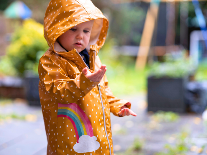 Young girl outdoors in a yellow raincoat with white polka dots and a rainbow on the pocket. The girl is looking at her hand