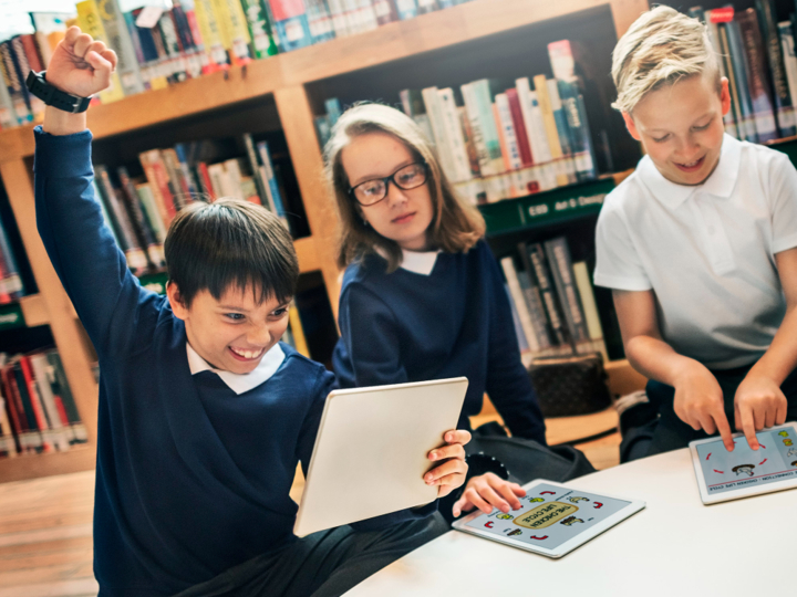 Three children in school uniform sitting in a class on ipads. One boy is raising his fit into the air with a happy and excited grin on his face