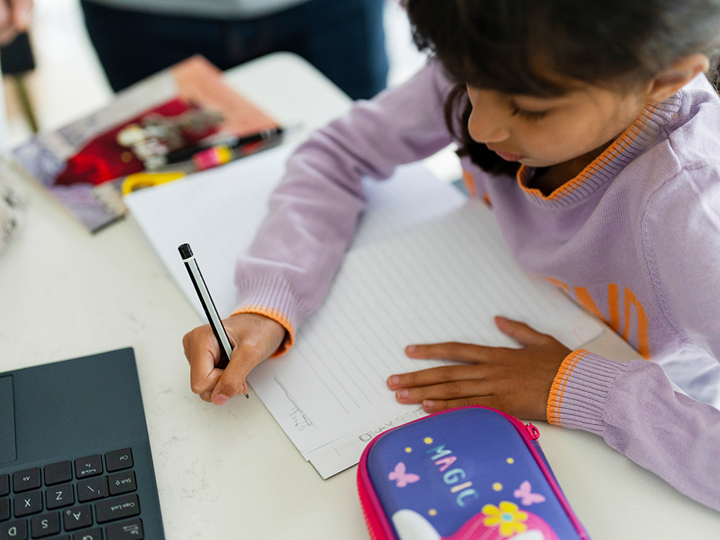 Girl writing on paper next to a space-themed pencil case and a laptop