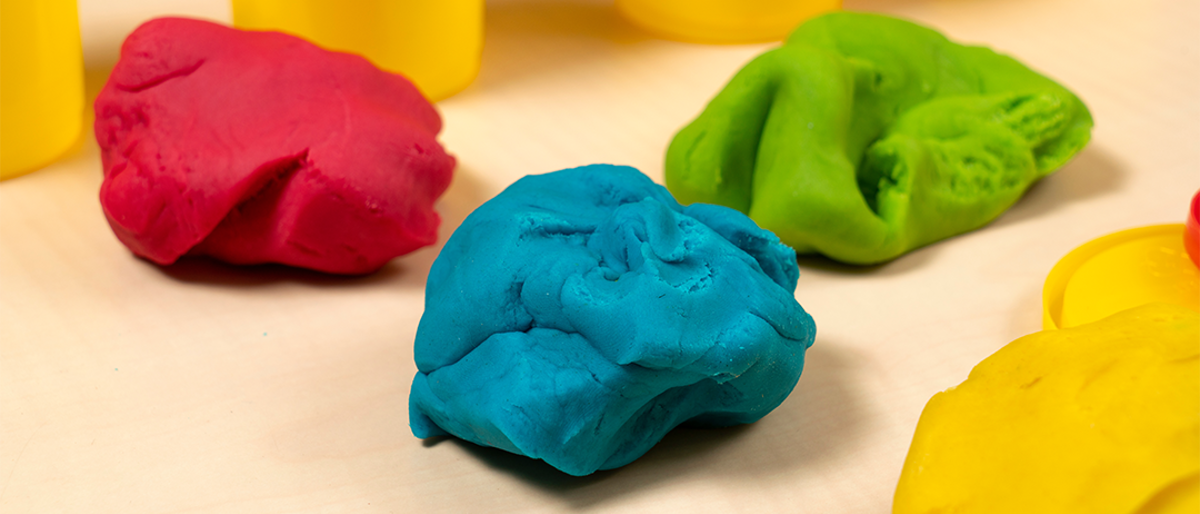 Red, blue and green lumps of playdough with yellow playdough pots in the background. They are all on a wooden table