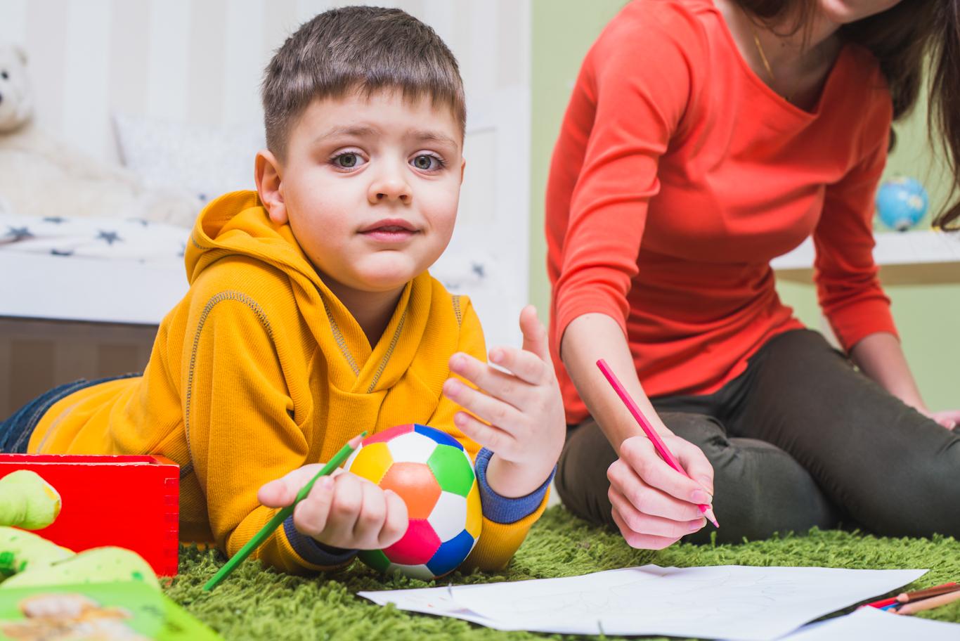 Boy in yellow hoodie lying on his stomach. He's got a small ball between his arms and a workbook in front of him. He is holding a pencil and looking at the camera like he's just been interrupted during the task.
