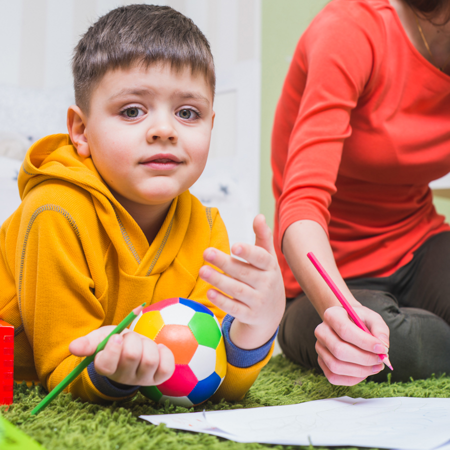 Boy in yellow hoodie lying on his stomach. He's got a small ball between his arms and a workbook in front of him. He is holding a pencil and looking at the camera like he's just been interrupted during the task.