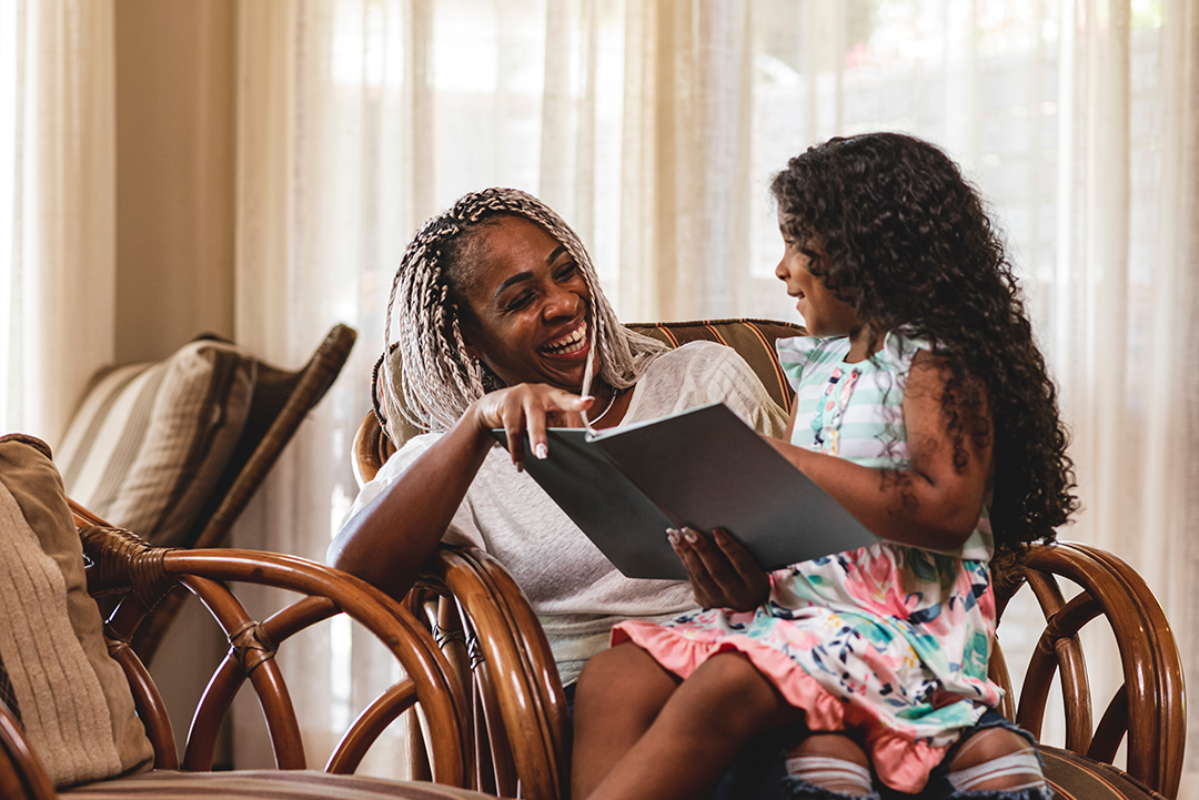 girl sitting on lap of laughing adult. Girl is smiling and reading book to adult