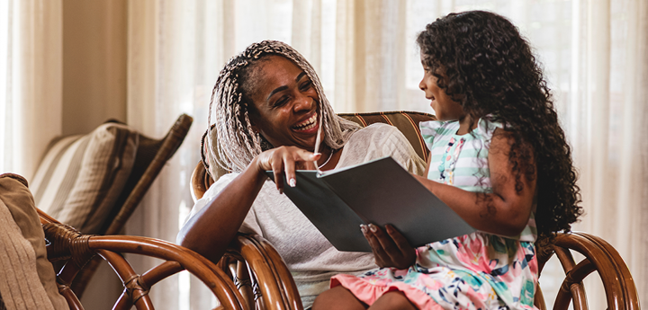 girl sitting on lap of laughing adult. Girl is smiling and reading book to adult