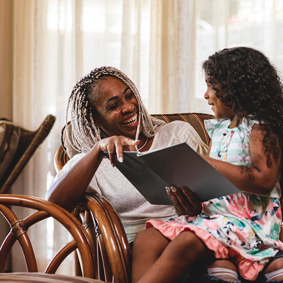 girl sitting on lap of laughing adult. Girl is smiling and reading book to adult