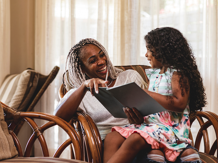 girl sitting on lap of laughing adult. Girl is smiling and reading book to adult
