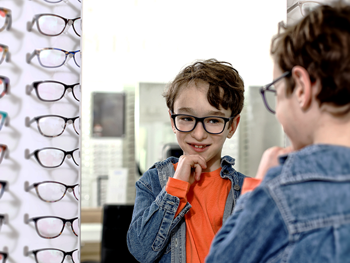 boy in orange long sleeve t-shirt and denim jacket admiring himself in the mirror at an opticians as he tries out new glasses