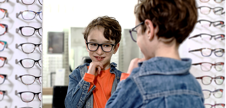 boy in orange long sleeve t-shirt and denim jacket admiring himself in the mirror at an opticians as he tries out new glasses