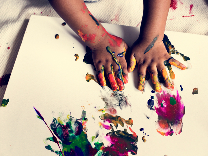 Young child's hands covered in different colour paints and pressing their hands against some pieces of paper.