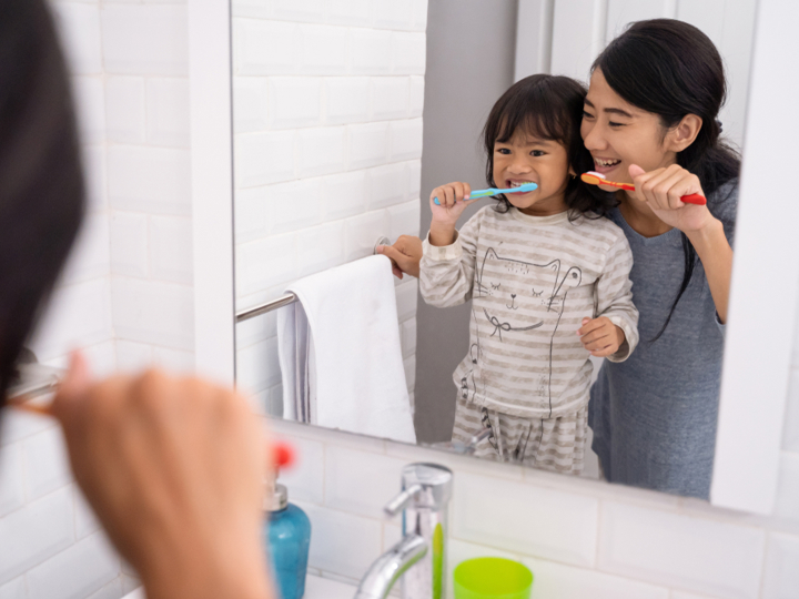 Mother and young girl standing in front of bathroom sink brushing their teeth and looking in the mirror.