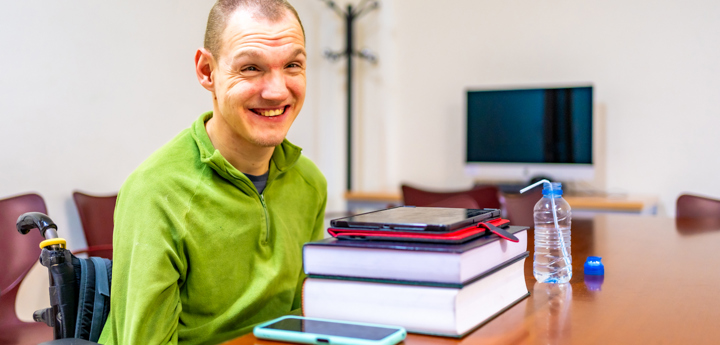 Disabled young adult sitting in a wheelchair at table with books a tablet a water bottle and a mobile phone