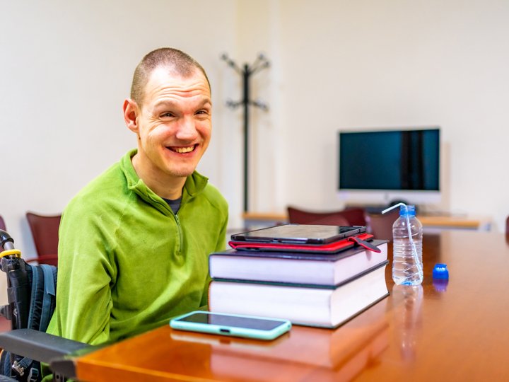 Disabled young adult sitting in a wheelchair at table with books a tablet a water bottle and a mobile phone