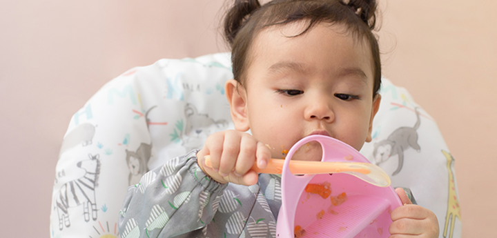 Baby in high chair playing with a pink bowl with orange spoon. Pieces of food are scattered across the highchair.