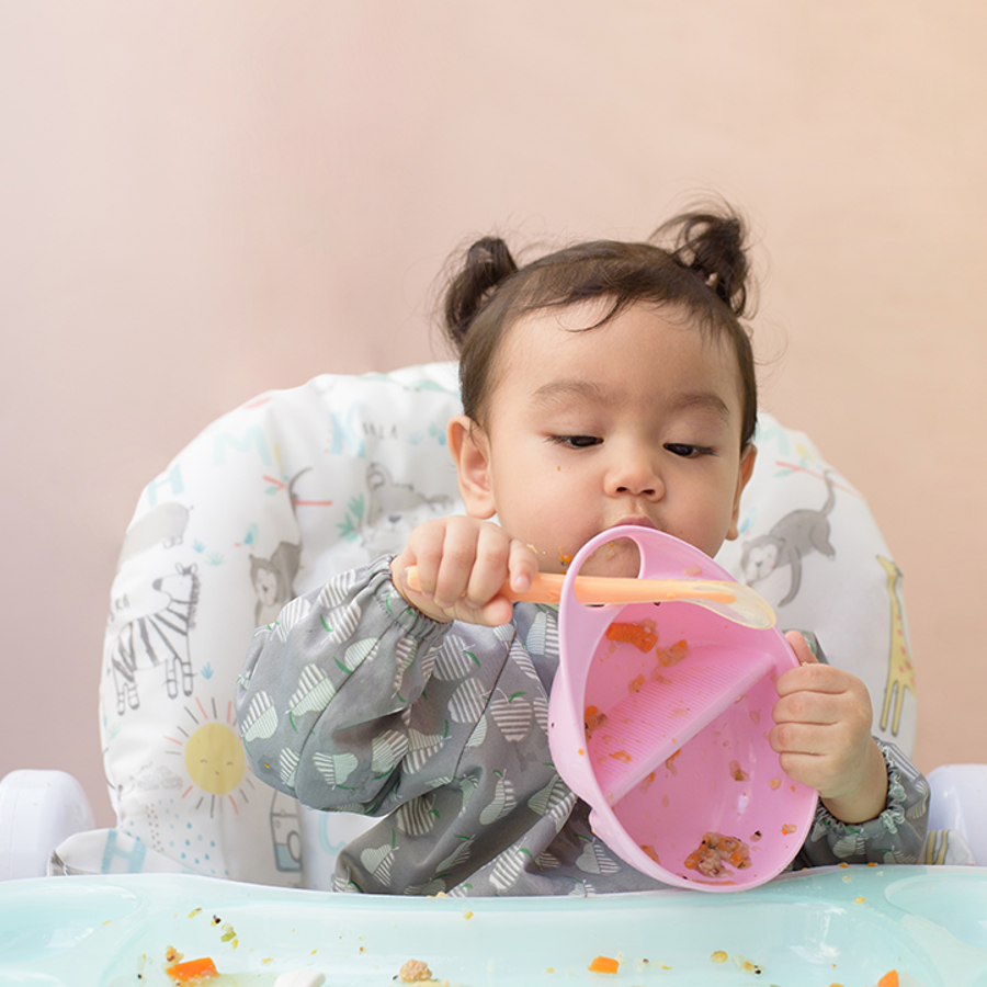 Baby in high chair playing with a pink bowl with orange spoon. Pieces of food are scattered across the highchair.