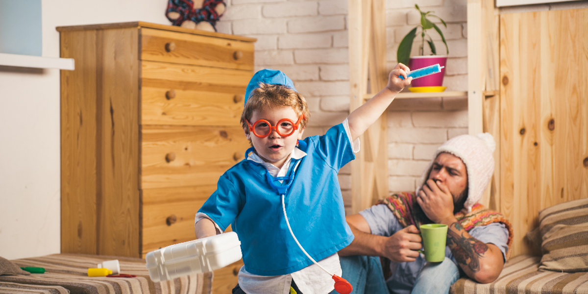 Child in nurses outfit with a toy stethoscope, toy syringe and a carrier box. Adult man in the background coughing and sat on the ground with a woolly hat and green mug 