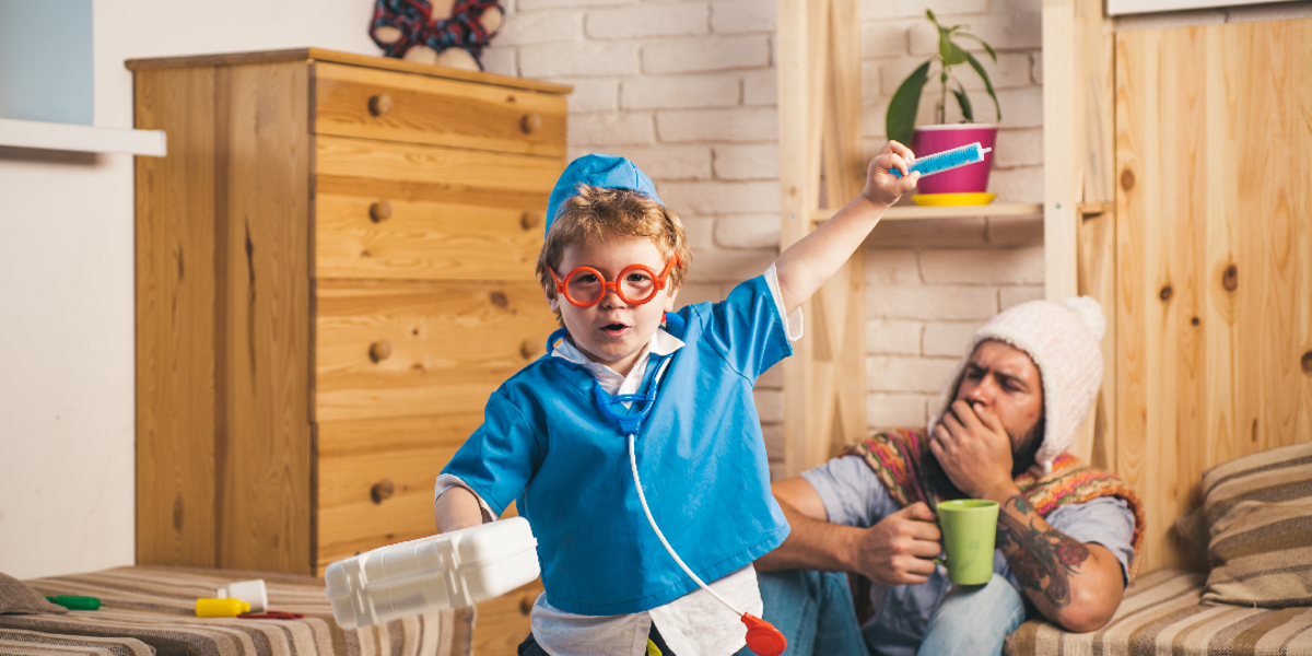 Child in nurses outfit with a toy stethoscope, toy syringe and a carrier box. Adult man in the background coughing and sat on the ground with a woolly hat and green mug