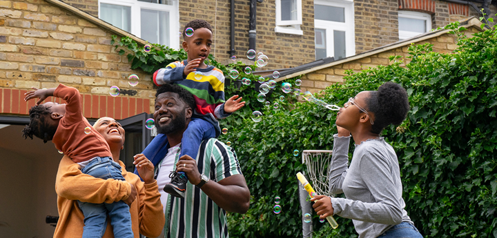 A family of standing in the garden. One girl is blowing bubbles and the dad is holding a young child on his shoulders whilst the mum is carrying another child. Both children are trying to grab the bubbles and everyone is laughing and smiling.